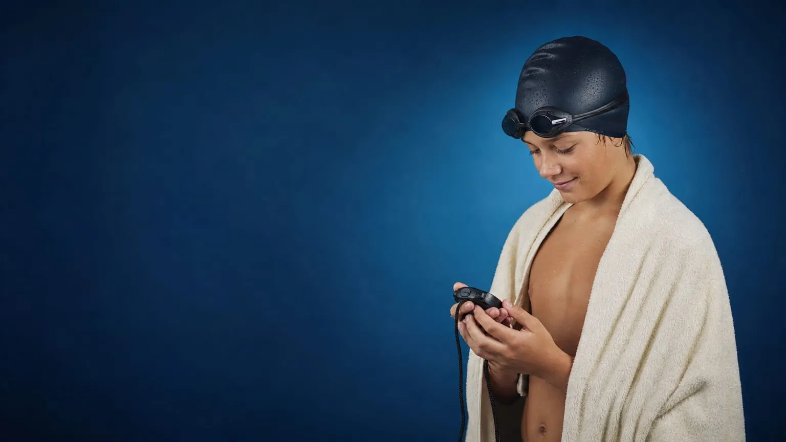 A young swimmer in a cap looking down at a handheld stopwatch held in both hands