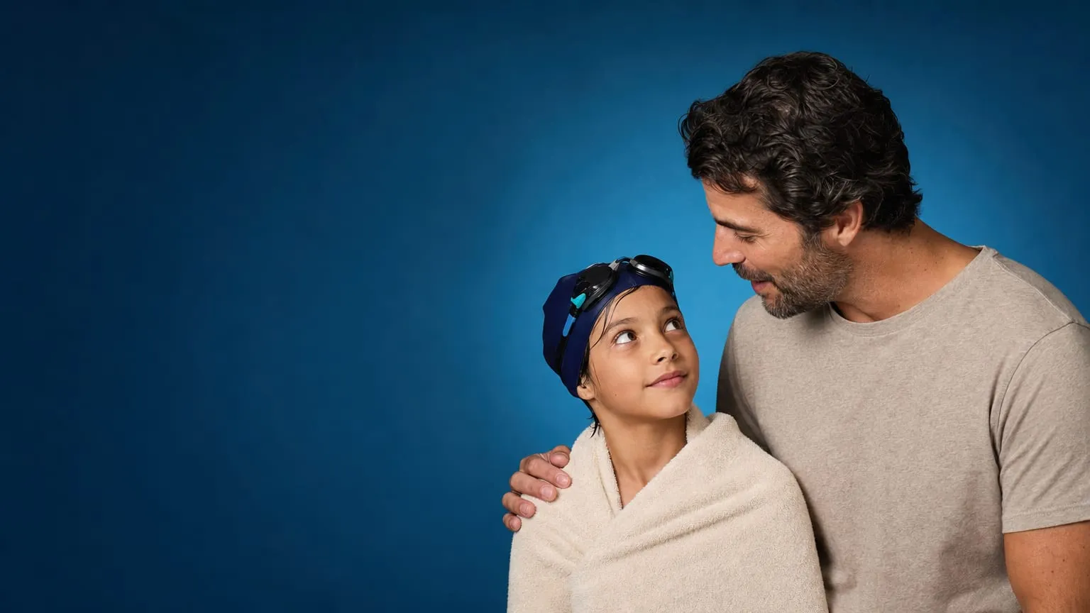 A parent leaning in mid-sentence beside their towel-wrapped young swimmer, who looks up listening