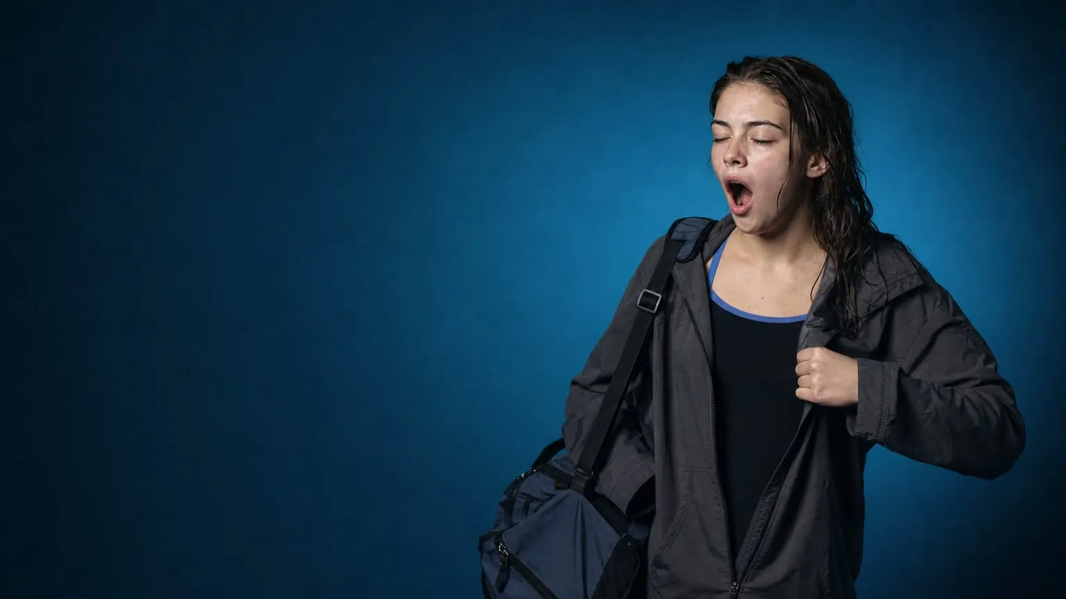 A tired teenage swimmer pulling on a jacket in the pre-dawn, a kit bag on one shoulder
