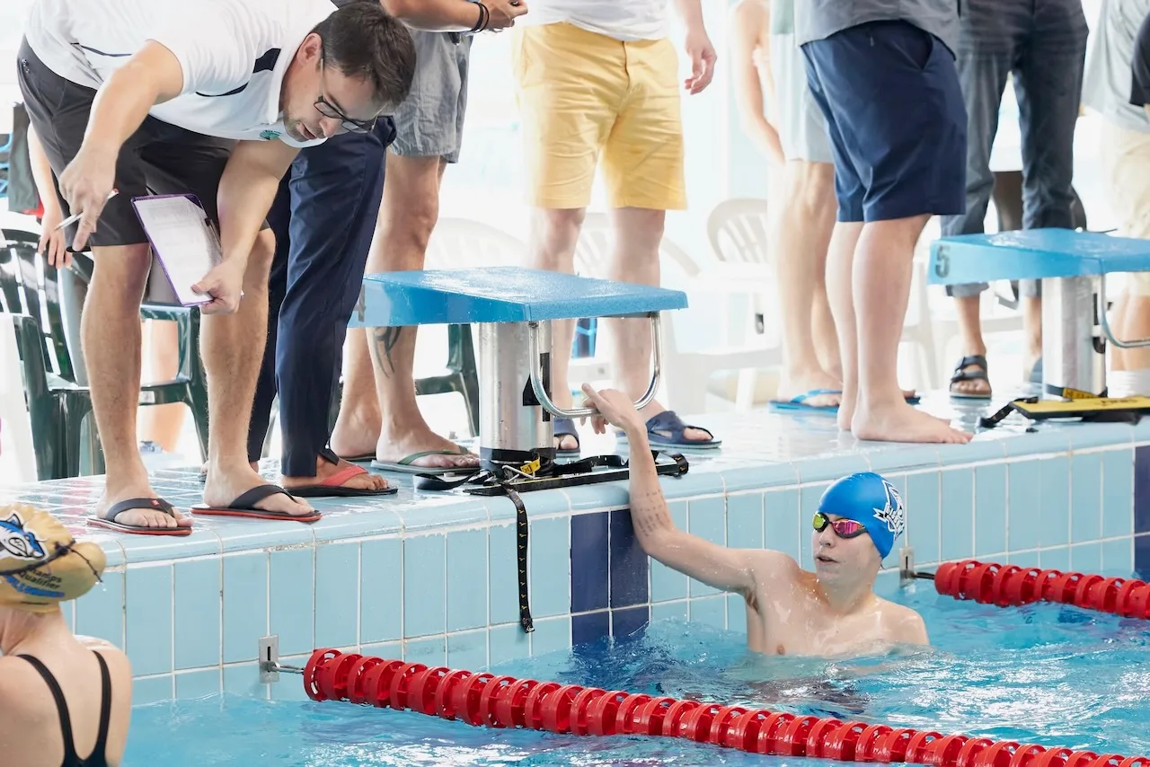 A young swimmer reaching up from the water to a coach leaning over the lane line, parents and teammates standing on the deck behind.