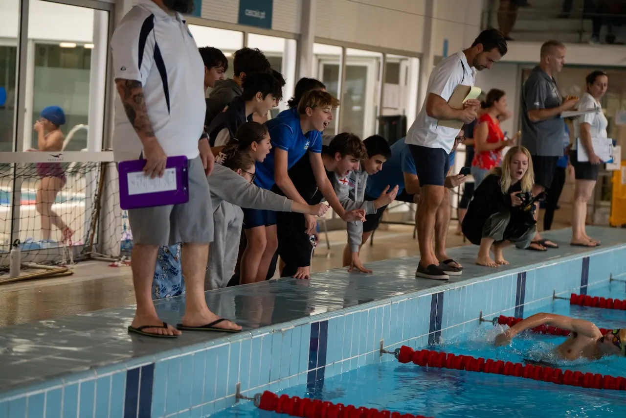 Colegas de equipa e um treinador a observarem um nadador a terminar, debruçados sobre a corda da pista