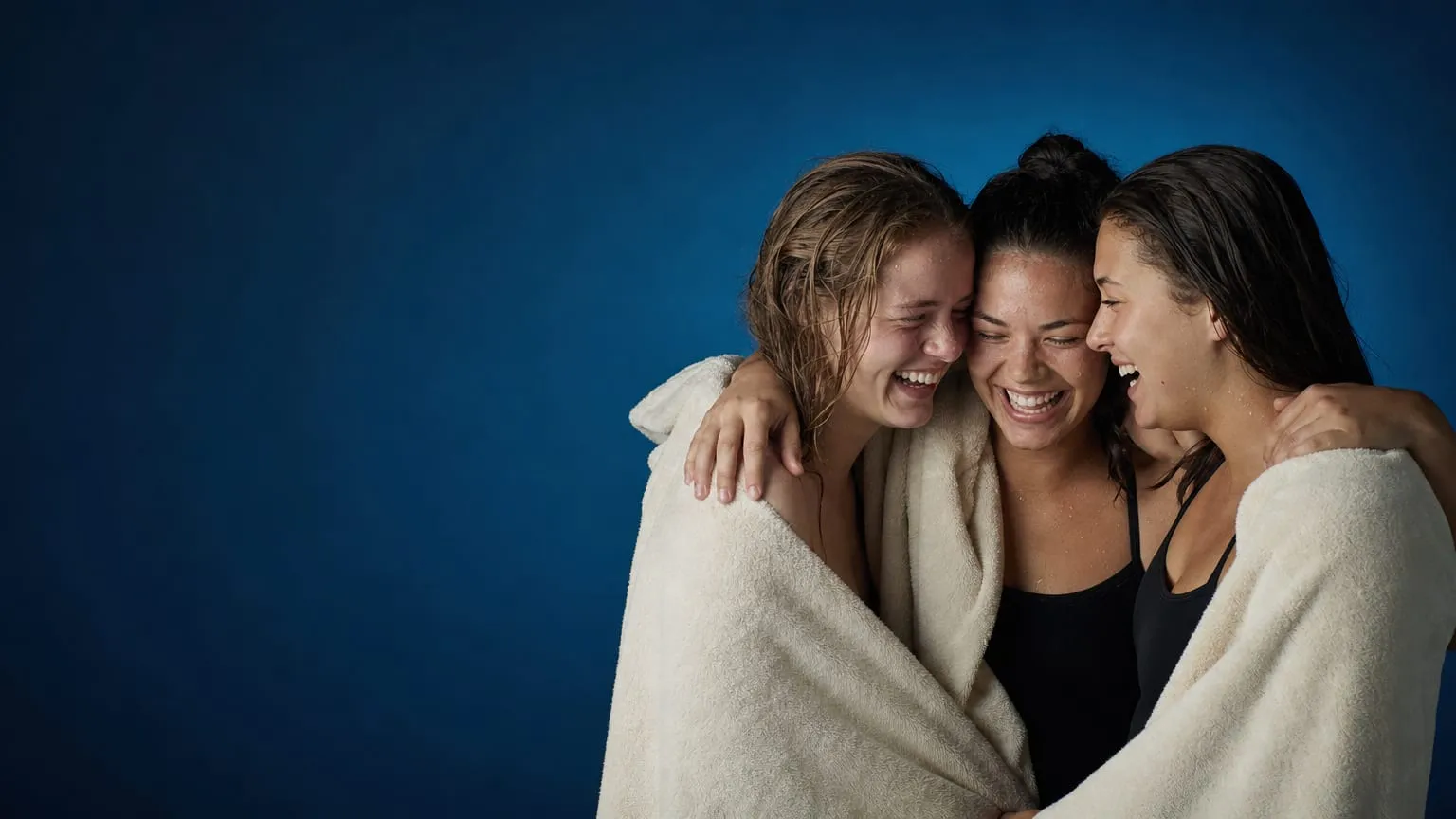 Three young teammates wrapped in towels with arms around each other's shoulders, laughing together