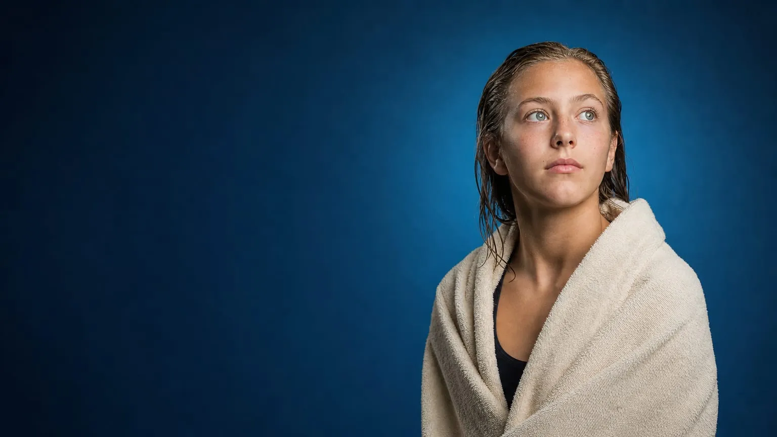 A teenage swimmer alone, wrapped in a towel, looking out in quiet thought